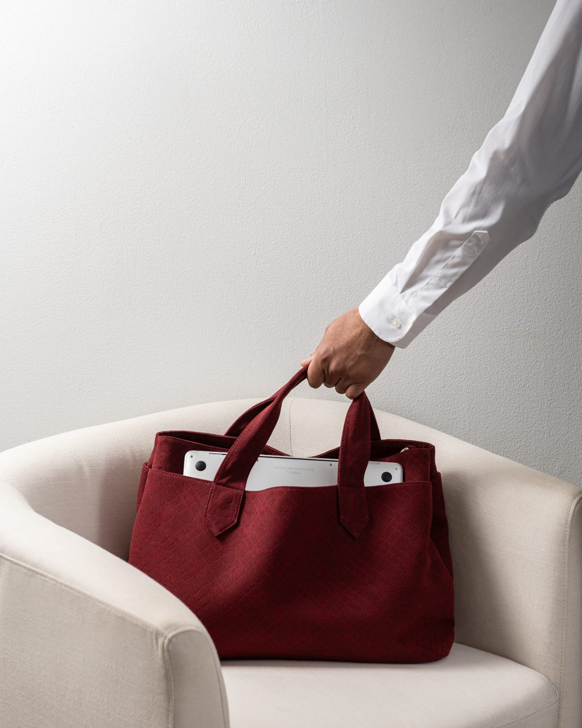 The Leon tote in Wine, lifted by its top handles from a cream armchair, a laptop visible inside.